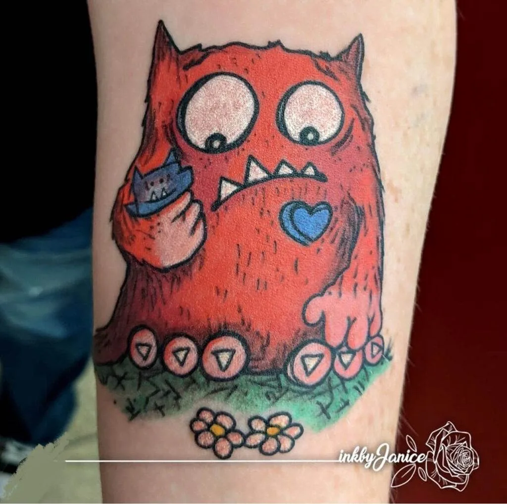 A tattoo of a red, furry monster with horns holding a smartphone in one hand and pressing buttons shaped like arrows with the other. It has a heart on its chest. The artist's signature reads "inkyJanice.