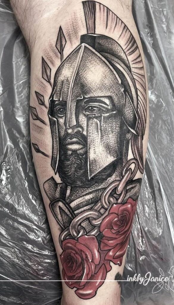 Tattoo of a warrior helmet with a bearded face, surrounded by chains and red roses, on a person's skin.